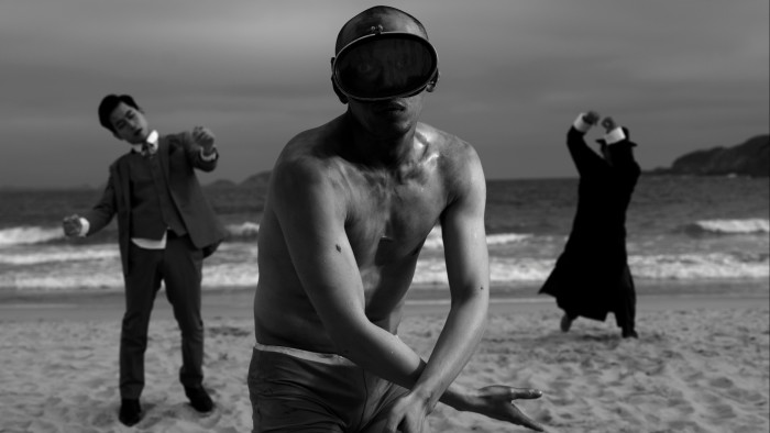 Black and white film still of a shirtless man on a beach with two men in suits in the background