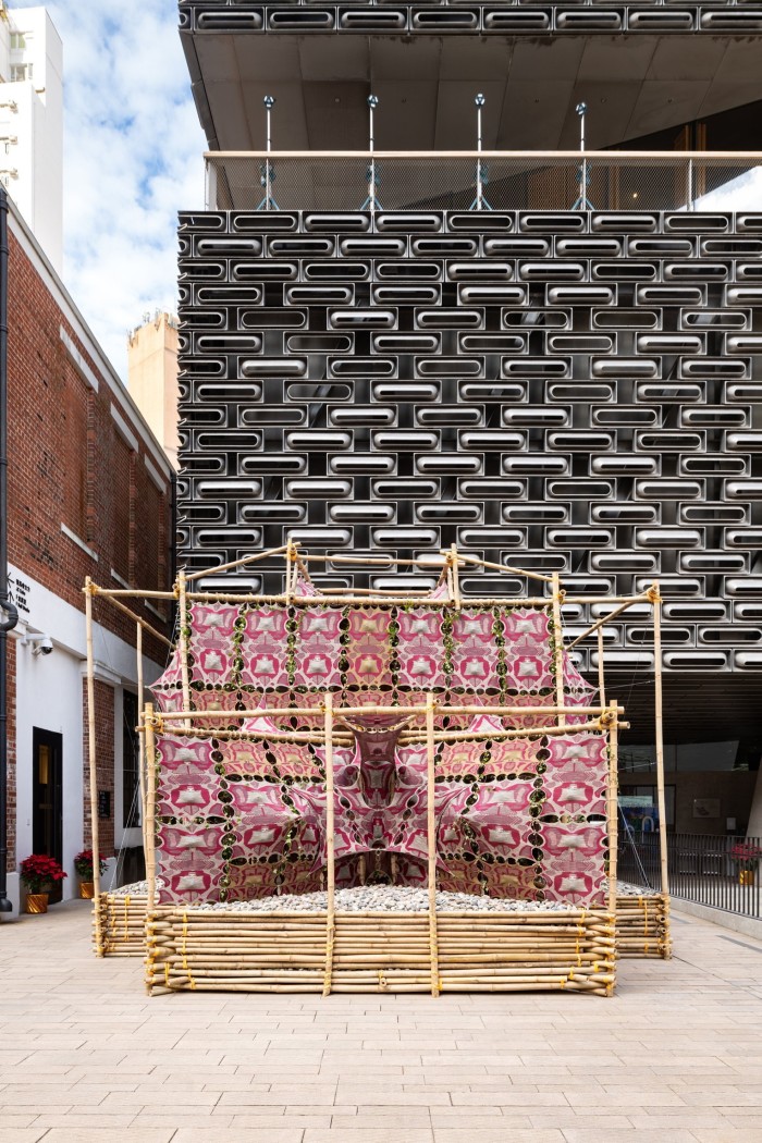 A small open bamboo shrine with pink banners sits in a courtyard before a sharp modern building