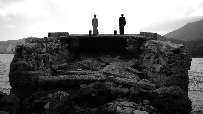 Black and white film still of two men in 1950s overcoats standing on a rocky ruin before the harbour