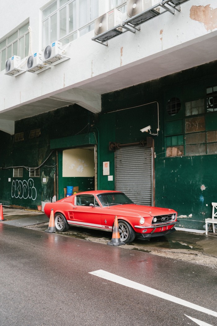 An old red Mustang sits outside a green industrial building
