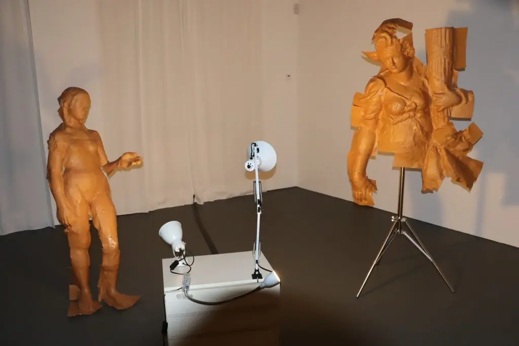 Two large orange-brown clay sculptures in a studio: a standing man on the left and a fragmented female figure on the right, with photography lights nearby.