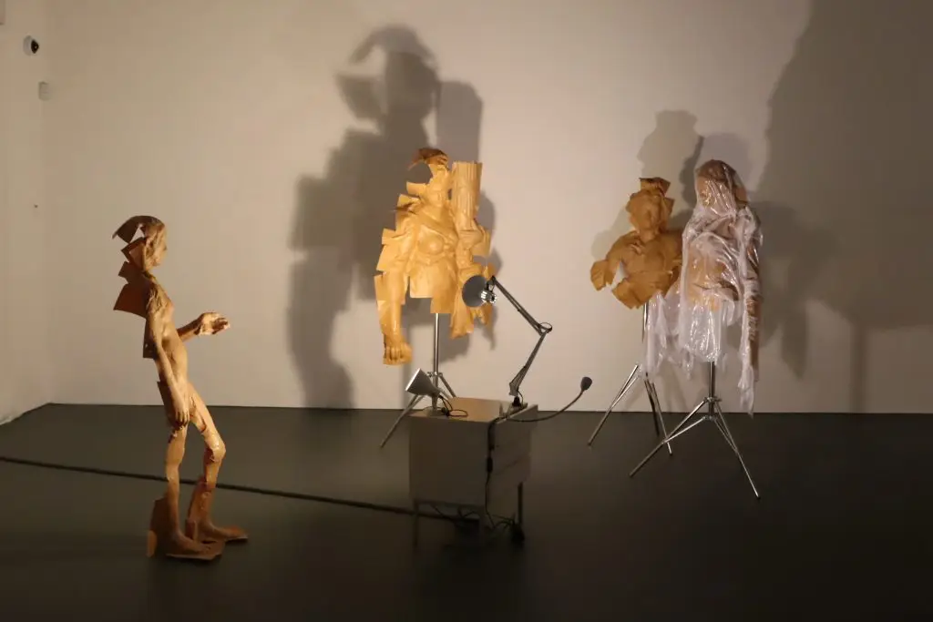 Art installation in a gallery: several brown paper sculptures on the floor and wall with microphones and stands in front of a pale wall.