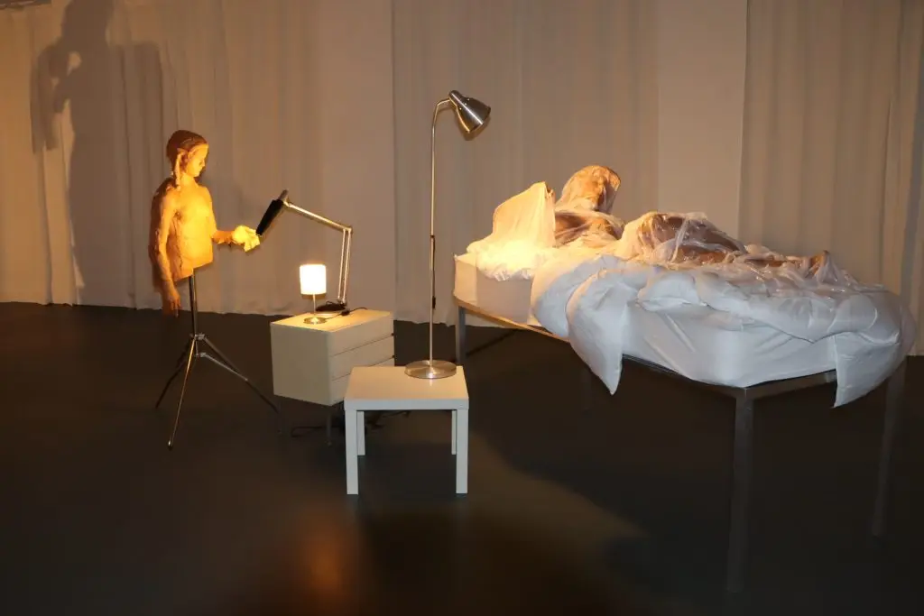 Dimly lit room with a hospital-style bed covered in clear plastic and a standing figure beside a desk lamp on a small table nearby.