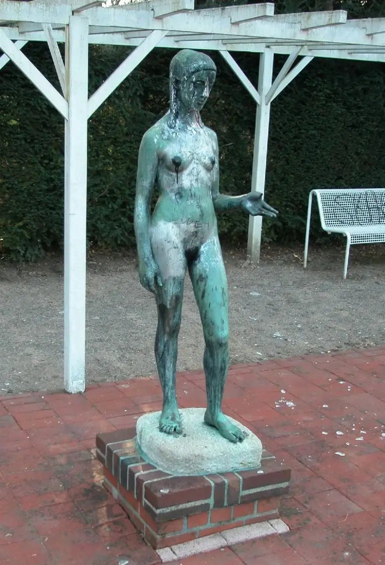 Bronze statue of a nude woman with a green patina, standing on a brick pedestal under a white wooden pergola.