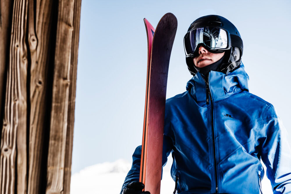A man in a blue jacket and helmet holding a pair of skis. J.Lindeberg Ski fall/winter 21. Campaign photographed by Axel Adolfsson.