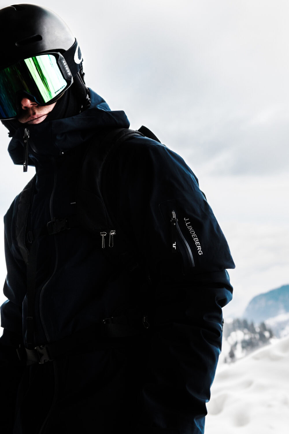A man wearing a helmet and a jacket standing on a snowy mountain. J.Lindeberg Ski fall/winter 21. Campaign photographed by Axel Adolfsson.