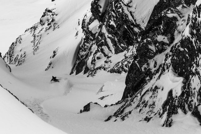 Andy Vogel in the steep in Engelberg. Snow-covered mountain landscape with a skier navigating steep terrain, rugged rocks, and pristine powder, captured in high-contrast black and white photography by Axel Adolfsson.