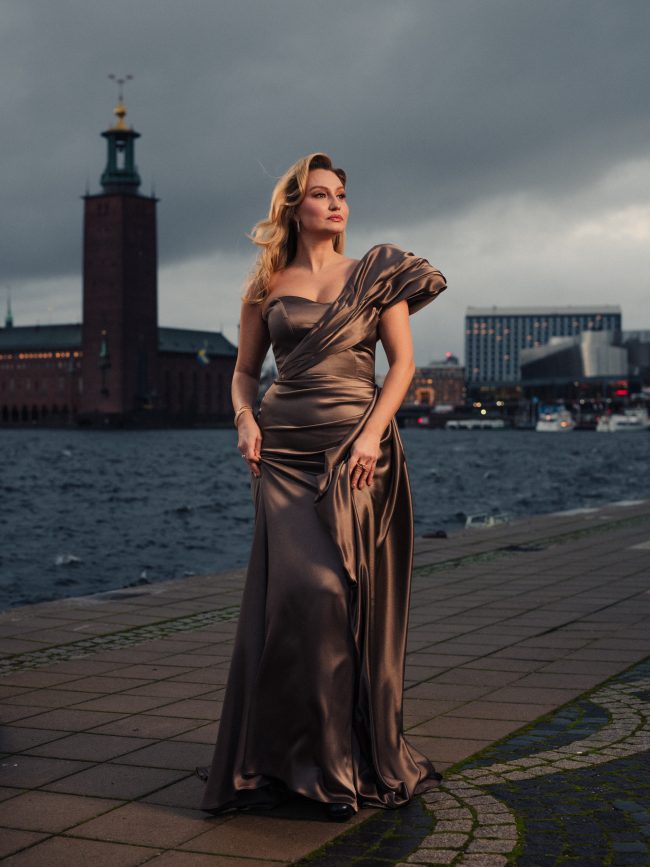 Elegant woman in glamorous metallic gown by the water with Stockholm Stadshus in the background, fashion photography. Ebba Busch photographed by Axel Adolfsson prior to Nobel 2025.