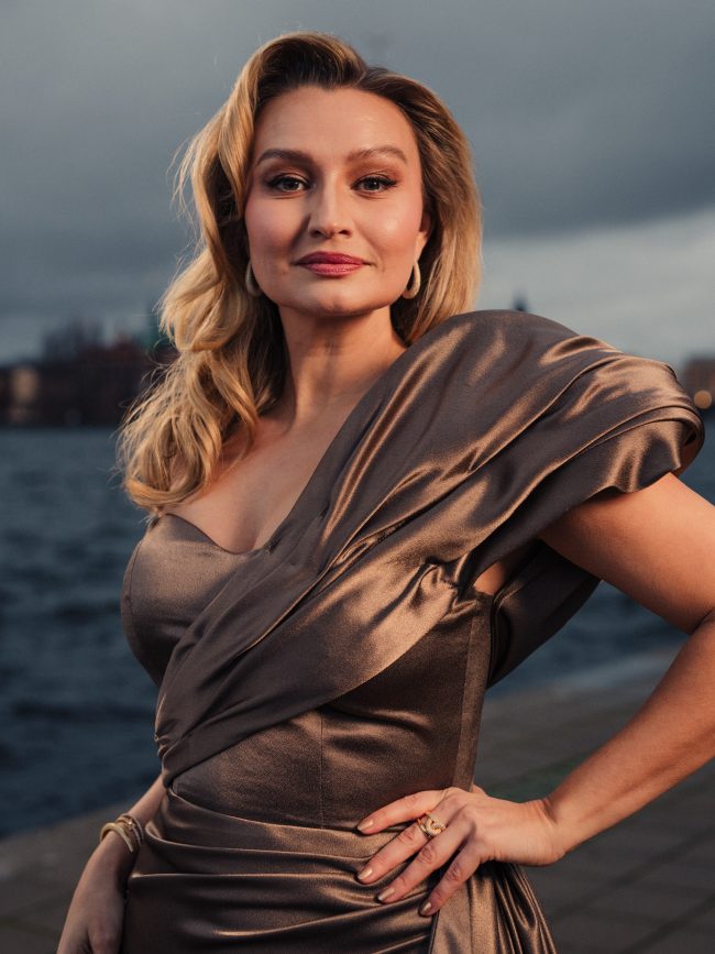 Elegant woman in satin dress by the waterfront during sunset. Ebba Busch photographed by Axel Adolfsson prior to Nobel 2025.