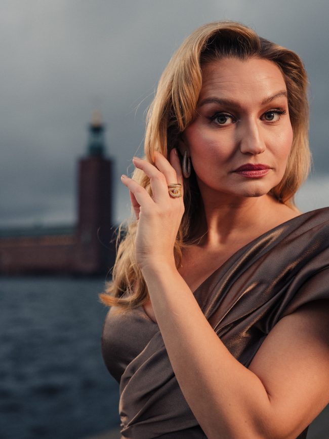 Elegant portrait of a blonde woman with jewellery, against Stockholm Stadshus backdrop. Ebba Busch photographed by Axel Adolfsson prior to Nobel 2025.