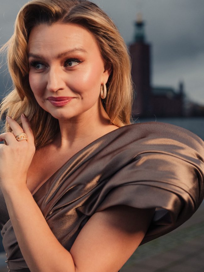 Elegant woman in metallic dress wearing jewellery, Stockholm Stadshus in background, fashion photography, evening light. Ebba Busch photographed by Axel Adolfsson prior to Nobel 2025.