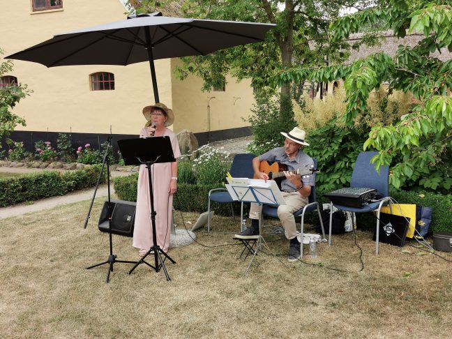 n dame synger og en mand spiller på guitar i en have under en parasol.