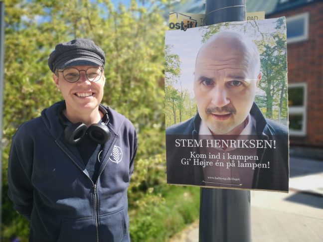 Mand med kasket står ved siden af valgplakat.