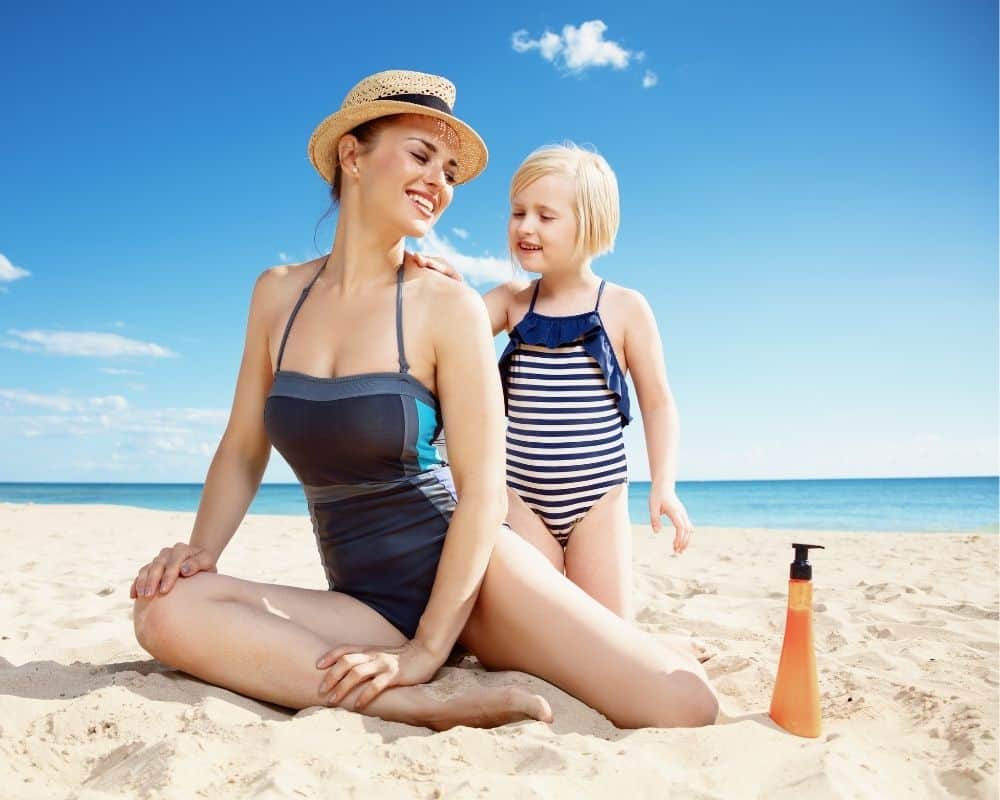 Mor og datter på stranden med solkrem i flasken ved siden av