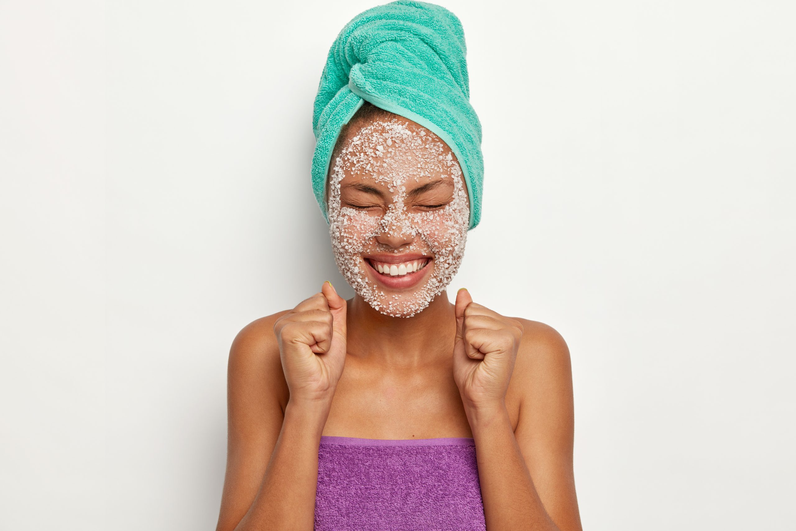 Horizontal shot of satisfied dark skinned woman clenches fists with pleasure, keeps eyes closed, applies sea salt mask, wrapped towel on head, isolated over white background. Body care concept