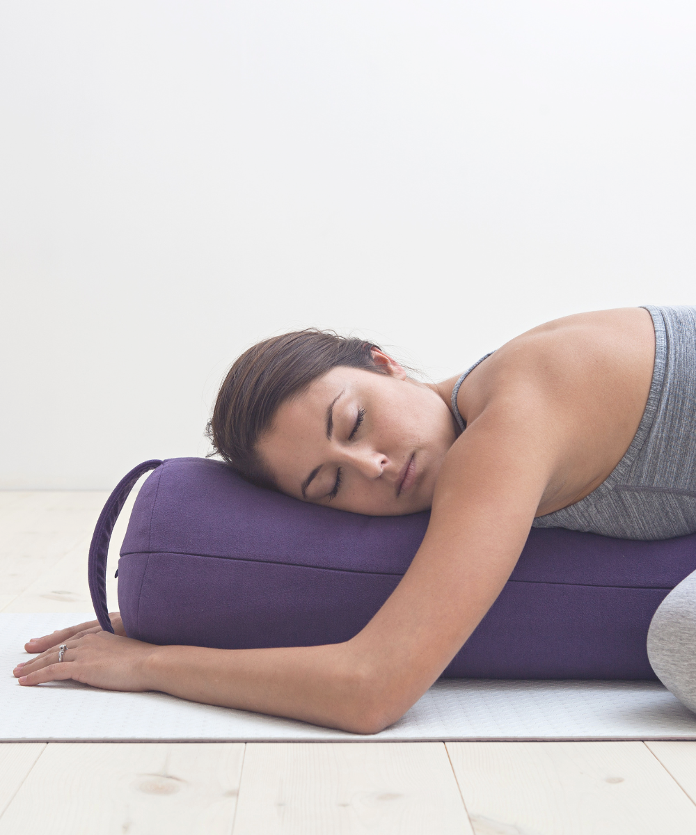 a woman resting in a restorative childs pose on a yoga mat. she has her eyes closed and is resting the side of her face on a purple yoga bolster. her forearms and elbows are resting on the yoga mat.