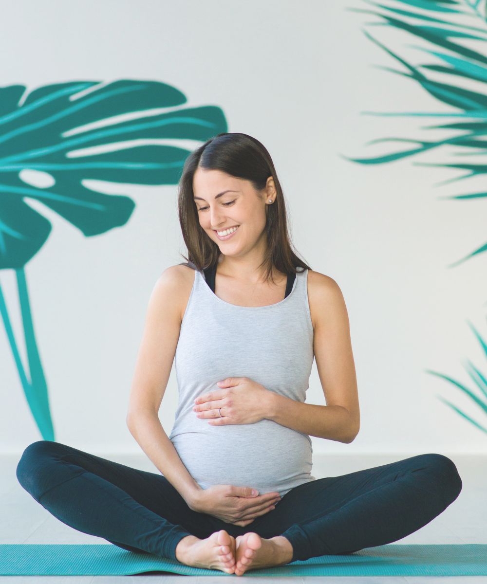 pregnant woman seated on a yoga mat practicing butterfly yoga pose for pregnancy yoga. she is smiling down and holding her pregnant belly.