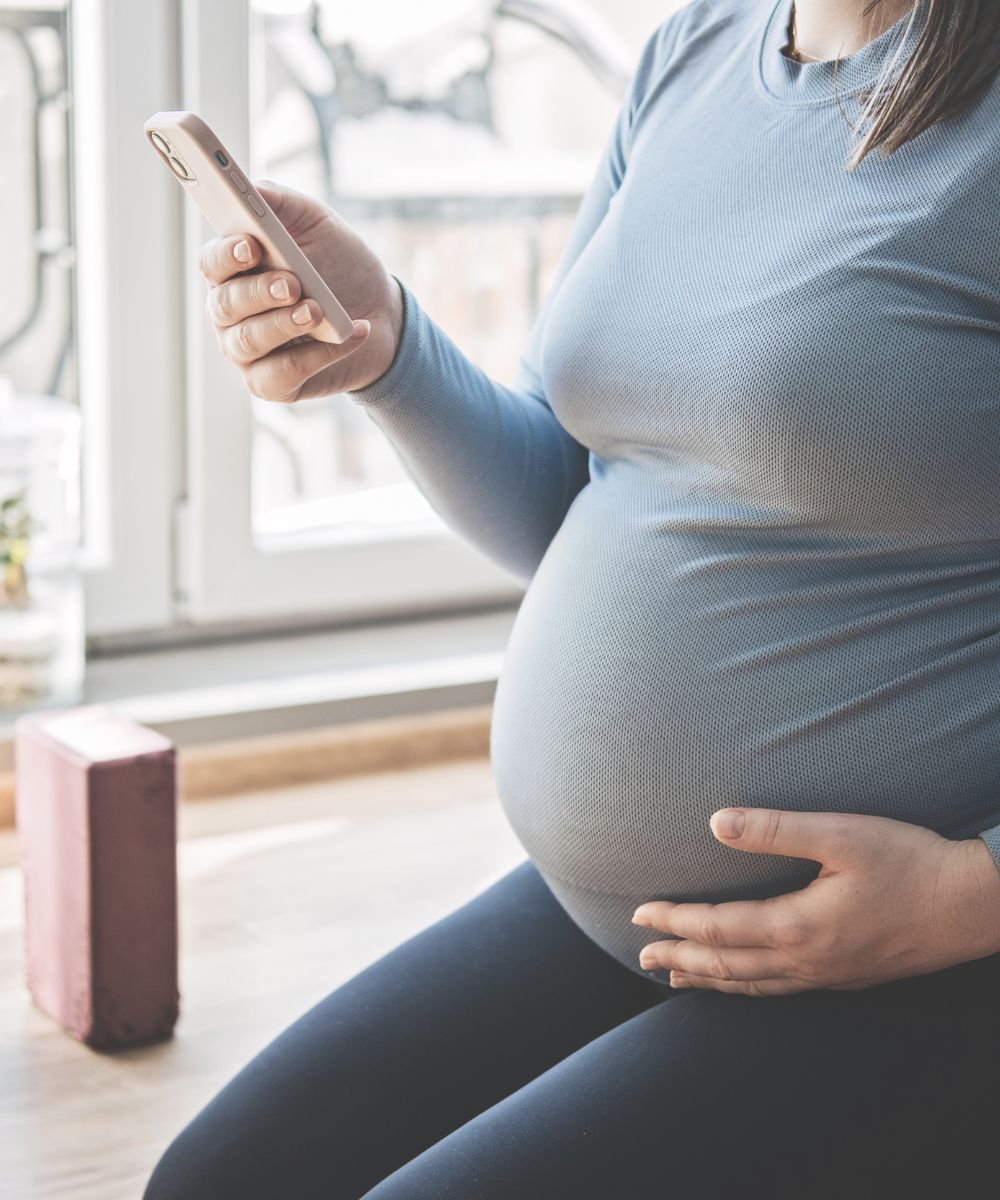 a pregnant woman sitting down in hero yoga pose. she is looking to her phone and practicing a digital pregnancy yoga program from home.