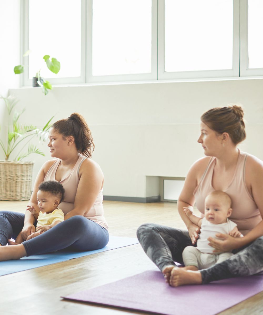 a mamma baby yoga class. two women are seated in butterfly yoga pose and holding their babies in their laps. the women are looking and listening to the yoga teacher and smiling. the babies look happy and content in the class.