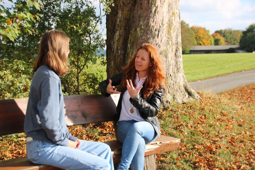 Corinna Zink sitzt im Park und führt im Rahmen ihres Angebotes für Persönlichkeitsentwicklung und Wohlbefinden ein Beratungsgespräch mit einer Frau, vielleicht als nächstes mit dir.