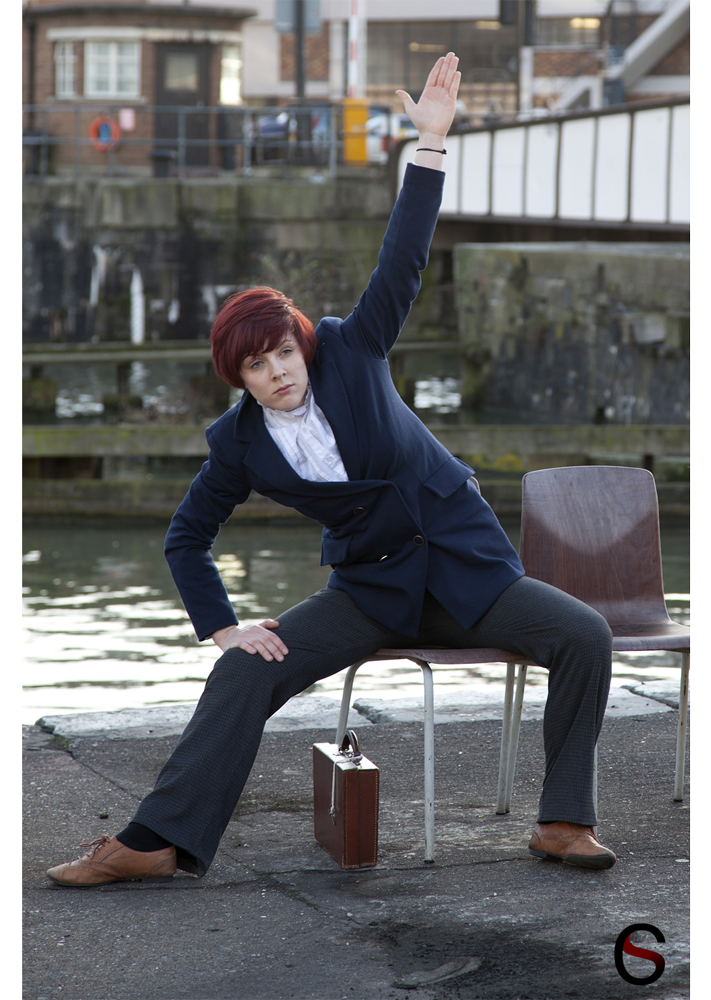 Woman in a navy blazer and white shirt posing on a chair by the waterfront, left arm raised, with a briefcase on the ground.