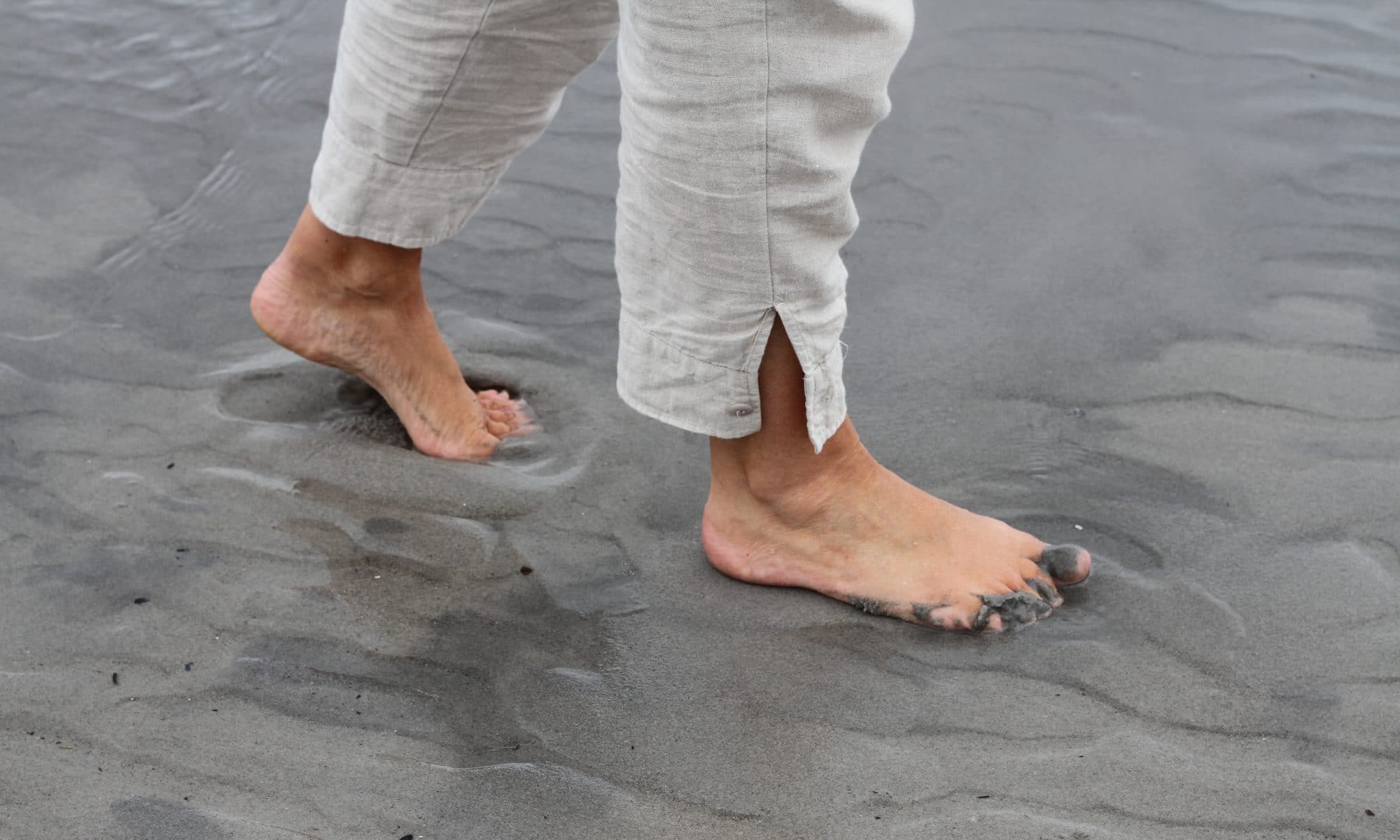 En person i et par lyse hørbukser, går barfodet på en våd sandstrand.