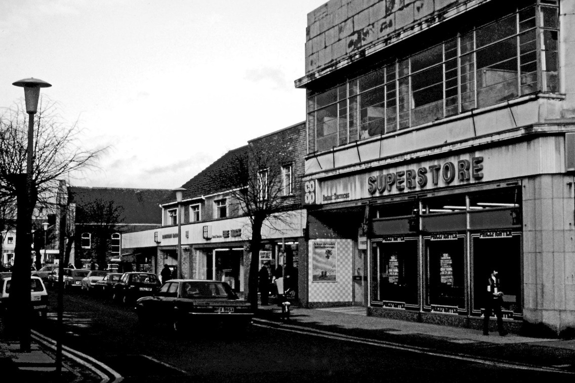 A black and white photo of a street with a car and buildings Description automatically generated with low confidence