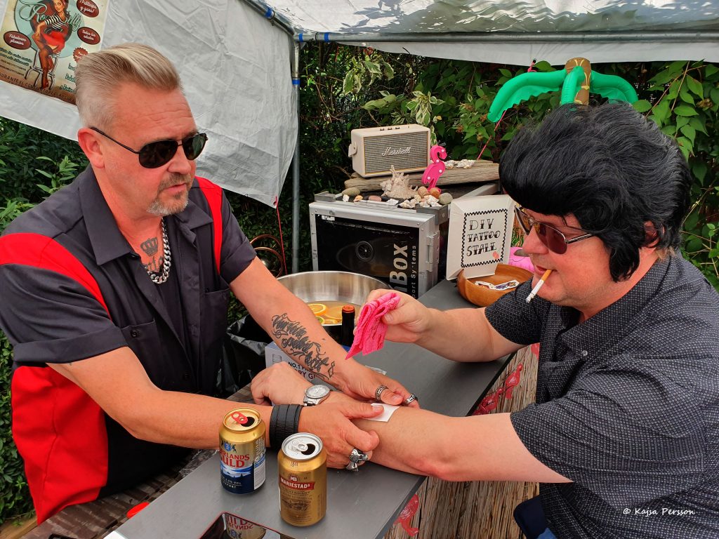 En kille hjälper en annan kille med Elvisfrisyr med en gnuggis tatuering