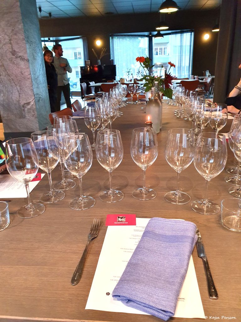 Uppdukat bord med vinglas för en vinprovning. Fem glas på varje plats med en beskrivning av kvällens viner och ostar framför och en blå tygservett