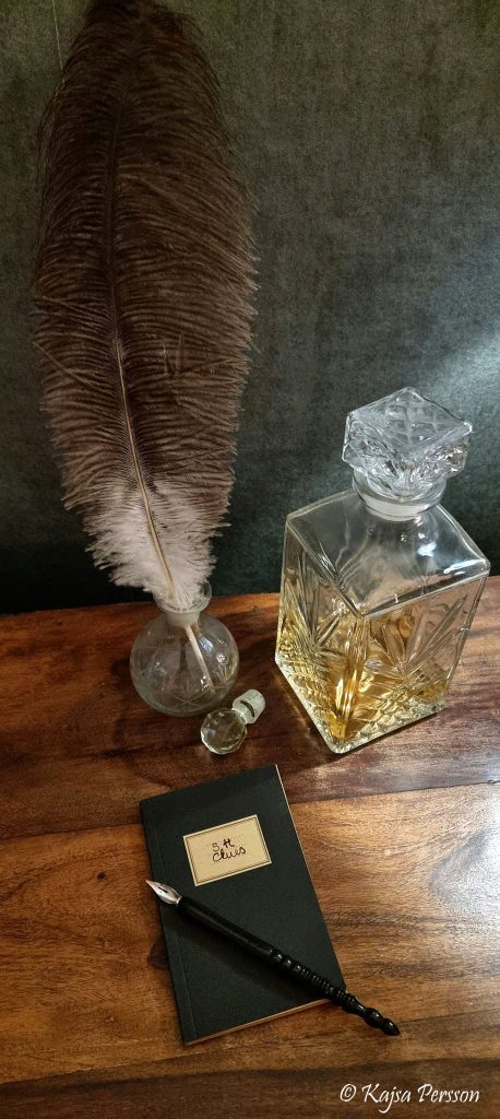 Anteckningsbok, fjäder och en karaff med Whiskey på en brun bänk