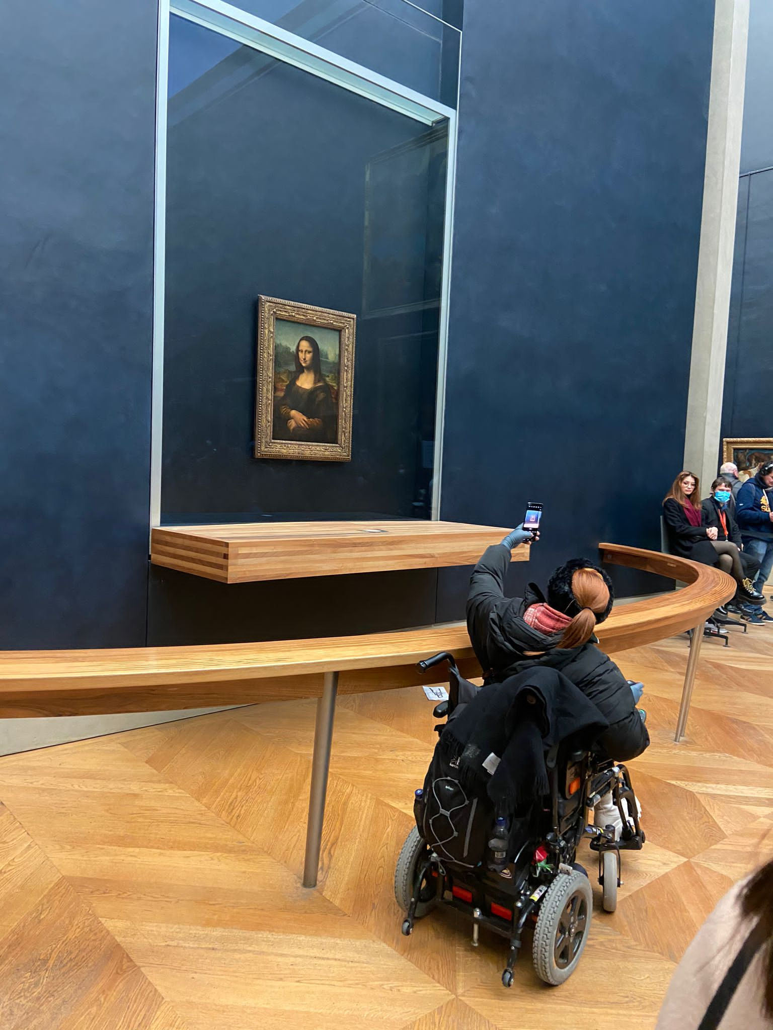 BLD experience setting in her wheelchair with her back and backpack facing the camera, she is holding up her phone taking a picture of the mona lisa