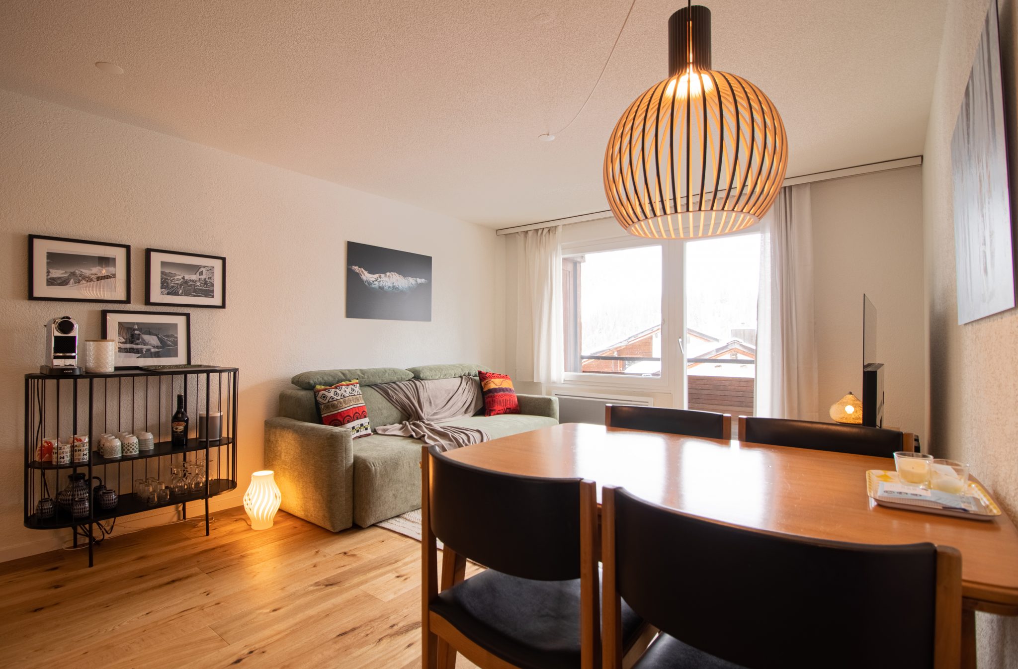 View of the living room of a modern airbnb studio with modern furniture and views of the snow-capped mountains. The decor is wood-toned and new. The property is located in Arosa.