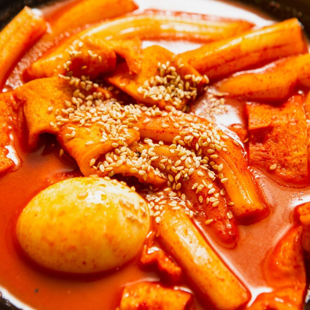 En bild på tteokbokki, koreanska risnudlar i en tjock, kryddig röd sås, toppad med kokt ägg och sesamfrön.