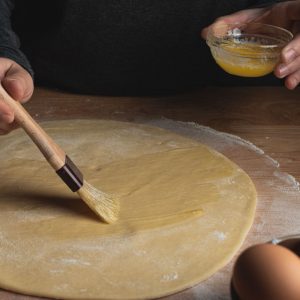 En person som varsamt penslar en deg med smält smör för att ge en gyllene och glansig yta vid gräddning. En person som varsamt penslar en deg med smält smör för att ge en gyllene och glansig yta vid gräddning.