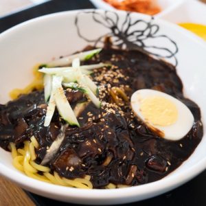 En skål med jjajjangmyeon, gula nudlar täckta med en rik svart bönsås och garnerat med ett halvt kokt ägg och gurka.