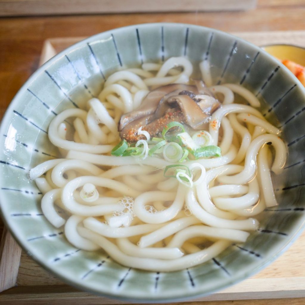 En skål med kokta udonnudlar i klar buljong, tjocka och mjuka med en len konsistens, garnerad med shiitake och salladslök.