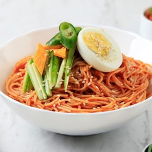 Bild på Bibimguksu, kalla, nudlar blandade i en röd, kryddig sås och toppade med gurka, sesamfrön och ett halvt kokt ägg