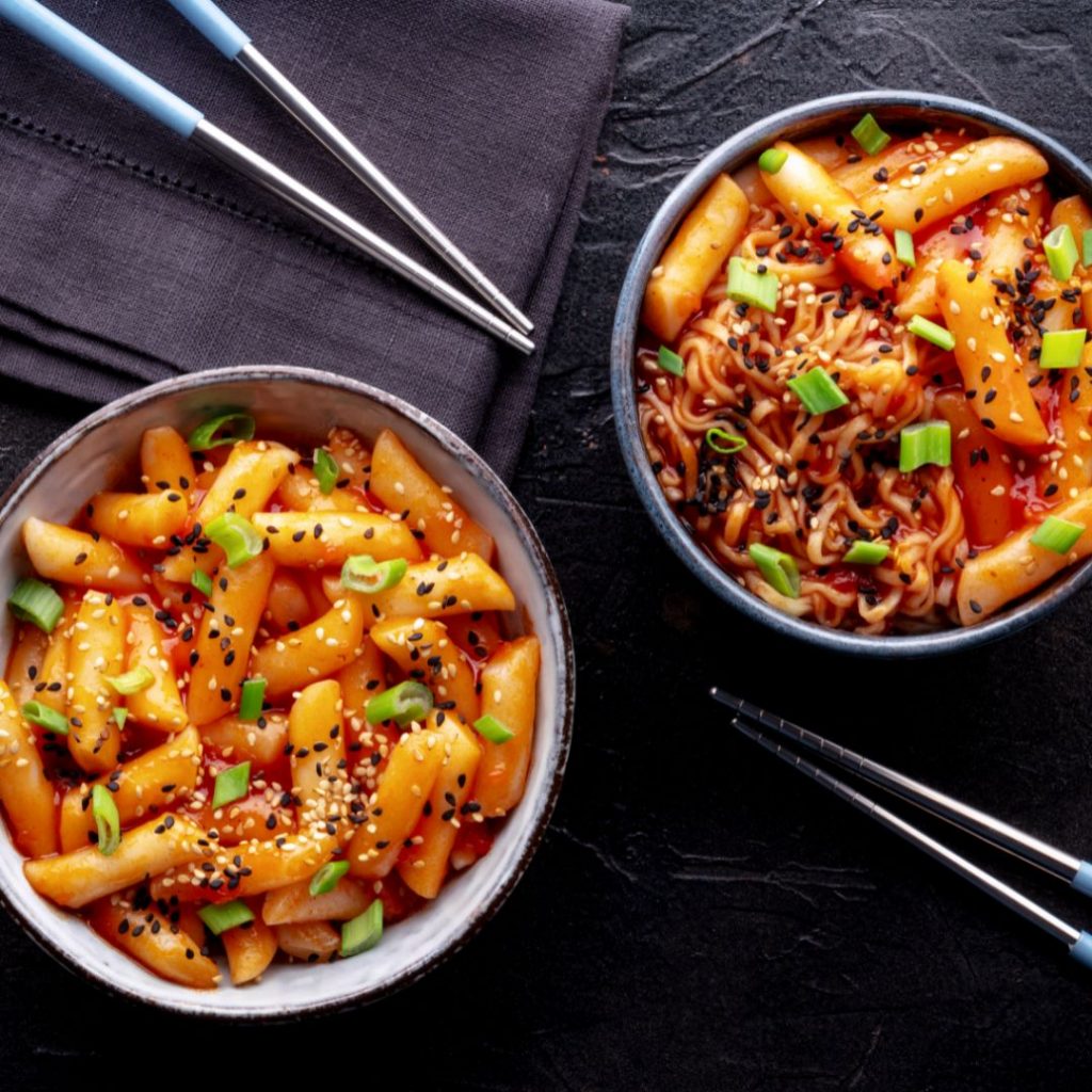 Två skålar med tteokbokki och rabokki, både med riskakor och i en het röd sås garnerade med salladslök och sesamfrön.