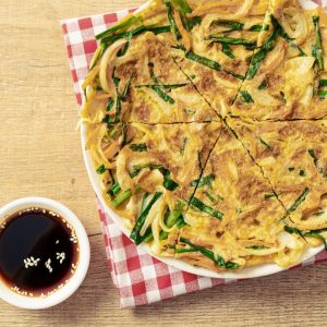 En stor pajeon, koreansk vårlökspannkaka, med en gyllenbrun yta med sojasås vid sidan.