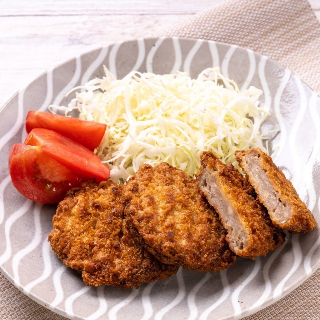 En tallrik med Menchi katsu, saftiga friterade köttfärsbiffar med krispigt yttre och strimlad vitkål och tomater vid sidan