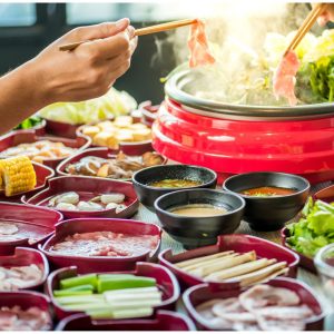 Ett bord med färska ingredienser och en rykande gryta med shabu-shabu, två händer med ätpinnar redo att doppa kött i grytan.