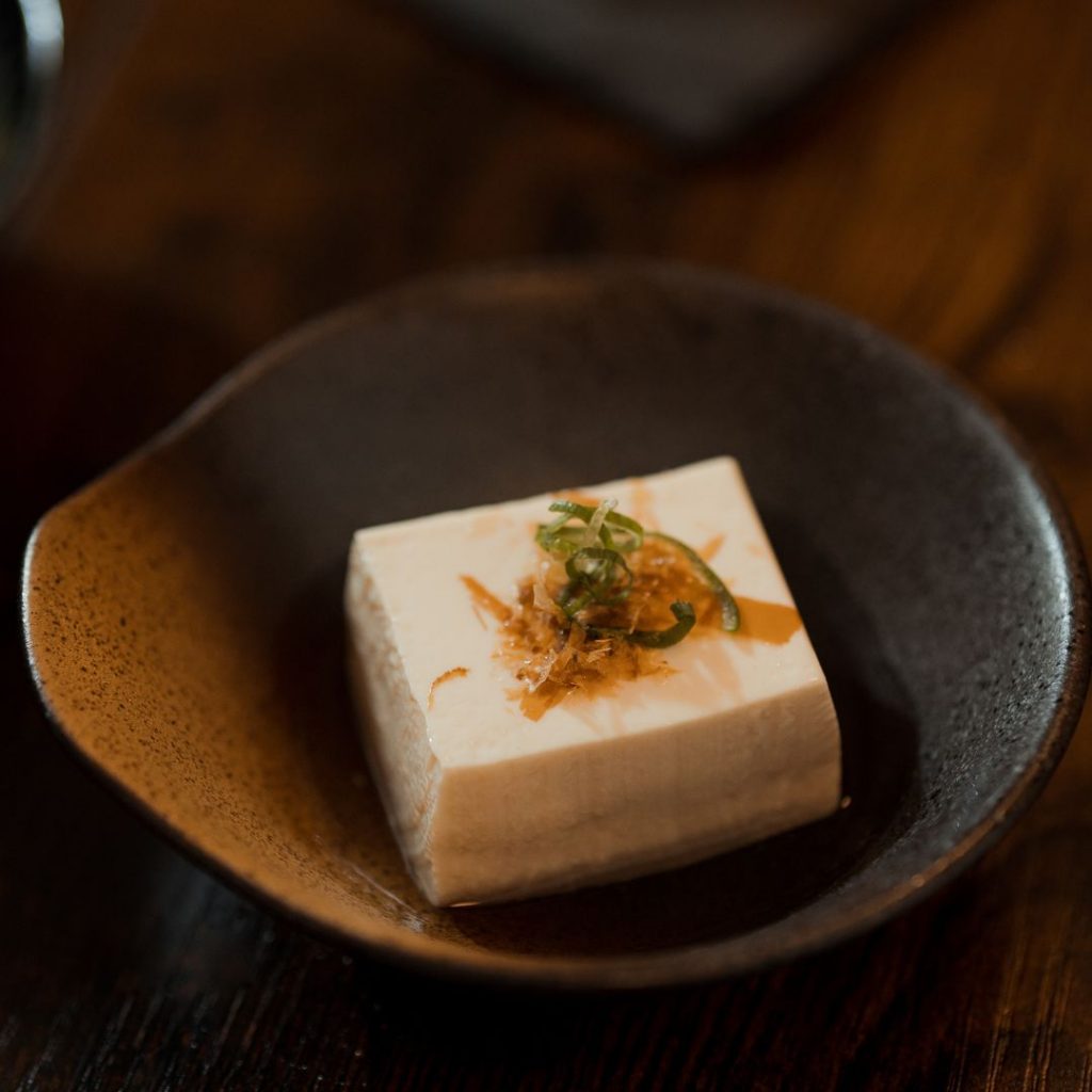 En skål med japansk yudofu, varsamt kokt tofu i en lätt dashi-buljong, serverad med sojasås och toppings.
