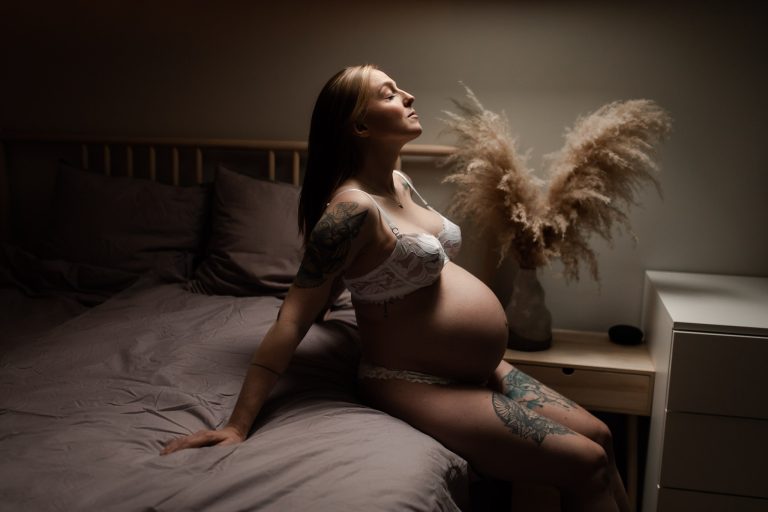 Gravid kvinna sitter på en säng iklädd endast underkläder med ljus som strålar ned mot hennes ansikte och mage, under en gravidboudoirfotografering i Stockholm