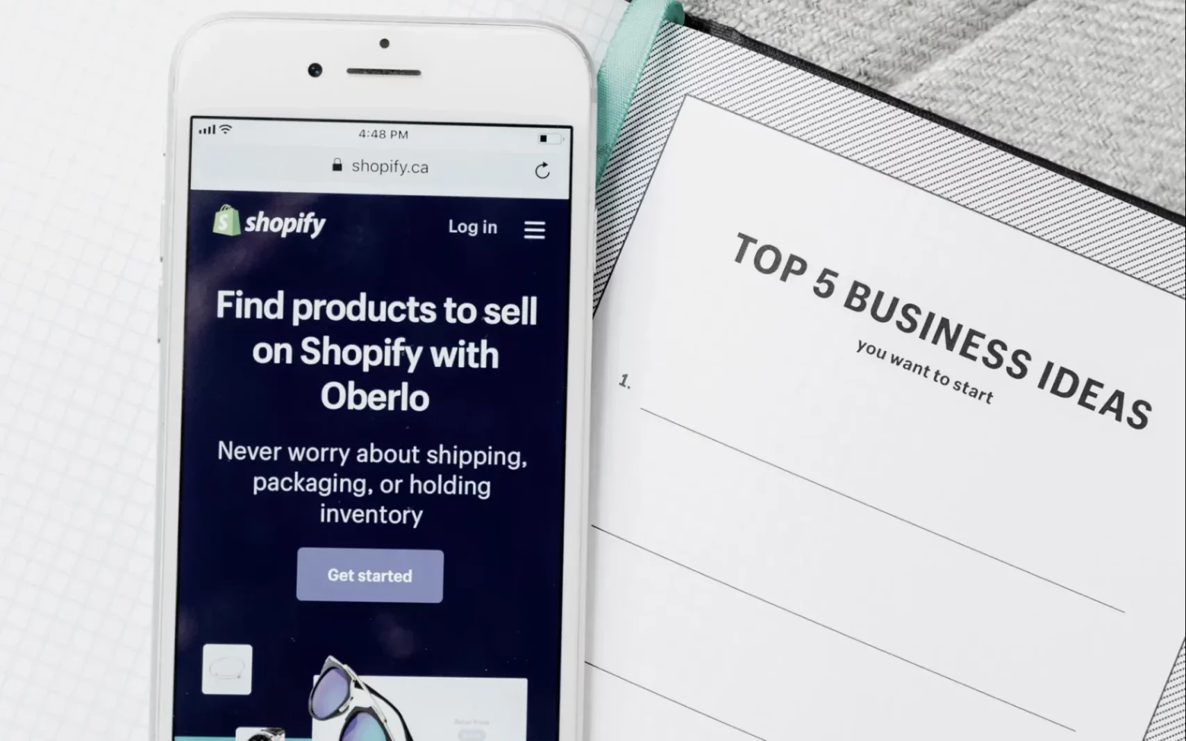 Smartphone mit geöffneter Shopify-Anwendung, das auf einem Notizblock mit der Aufschrift "Top 5 Business Ideas" liegt