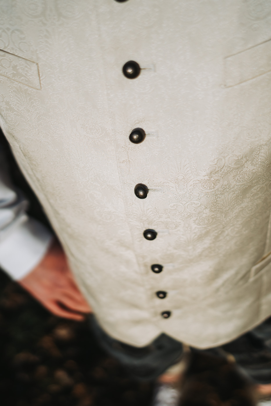 Detailaufnahme der eleganten Weste eines Bräutigams mit feinen Mustern und hochwertigen Stoffen. Die Weste ist sorgfältig über einem weißen Hemd getragen, mit einem sichtbaren Teil der Krawatte oder Fliege. Der Fokus liegt auf den Details der Weste, einschließlich der Knöpfe und des Stoffmusters.