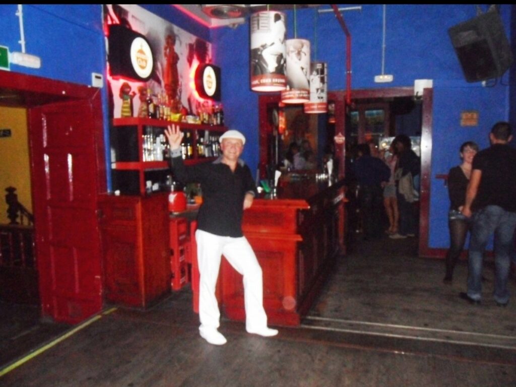 DJ Latino lehnt an einer Bar und winkt zur Kamera
