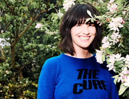 Head shot of writer Harriet Gibsone, standing in front of trees, wearing blue jumper with The Cure written on it in black