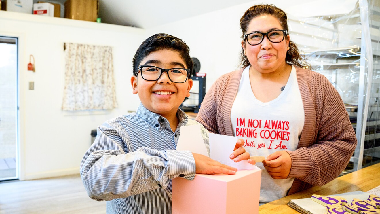 A photo of a baker standing next who her child who is holding a box that he is folding to place cookies in.