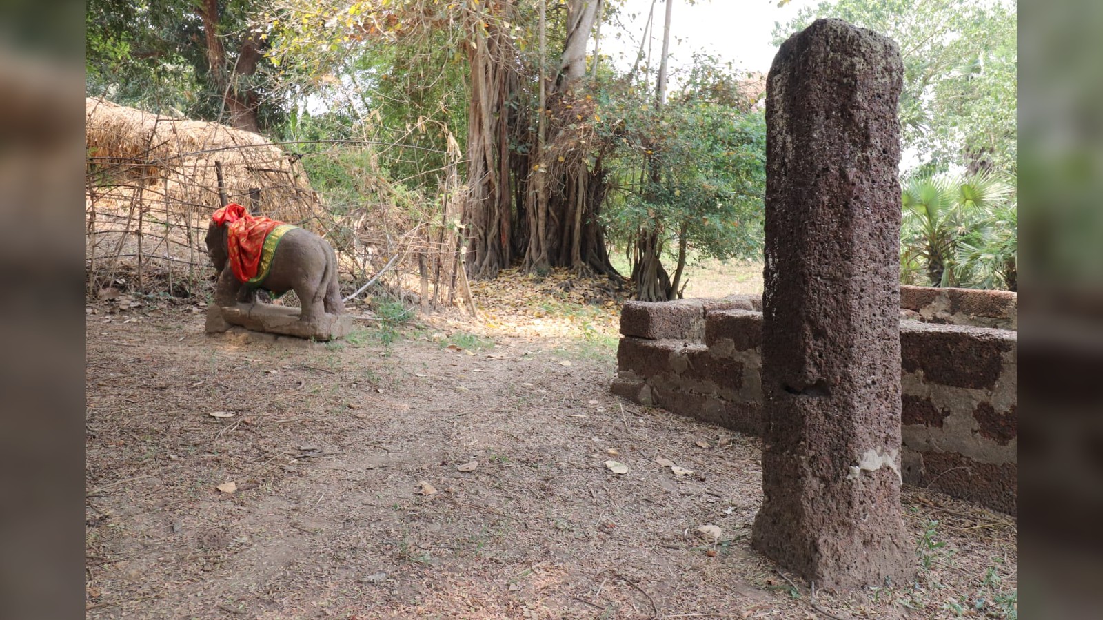 A reddish pillar is in the foreground with the elephant (partially covered with a red cloth) in the background in the field.