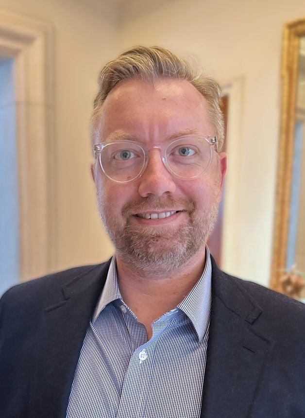 A blond man with a short hair and a graying beard wears clear-framed glasses, a blue checked shirt, and a blue suit jacket and smiles at the camera in a headshot.